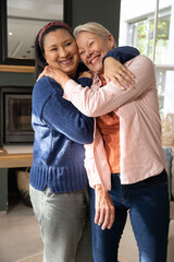 Naklejka premium Mature female friends hugging in living room, wearing sweater and jeans, near fireplace and window