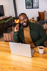 Naklejka premium African American green-shirted man talking on phone while working on laptop at table in living room