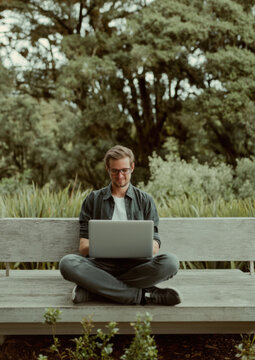 Remote work lifestyle digital nomad working on laptop outdoors. Young man sits on wooden bench surrounded by lush New Zealand native bush, combining business travel with nature tourism.