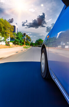 title: Low Angle Motion Blur of Blue Car Driving on City Street under a bright, sunlit sky with dramatic clouds.