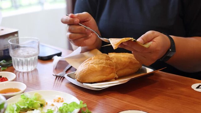 Close up video of unknown person using a fork to separate a piece of steamed flat cake from its banana leaf wrapping with fresh bread rolls on a wooden table,
