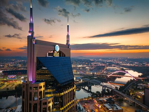  Aerial drone view of downtown Nashville skyline at sunset featuring the iconic AT&T &lsquo;Batman&rsquo; Building, Cumberland River, and illuminated city bridges with vibrant evening colors.&rdquo;