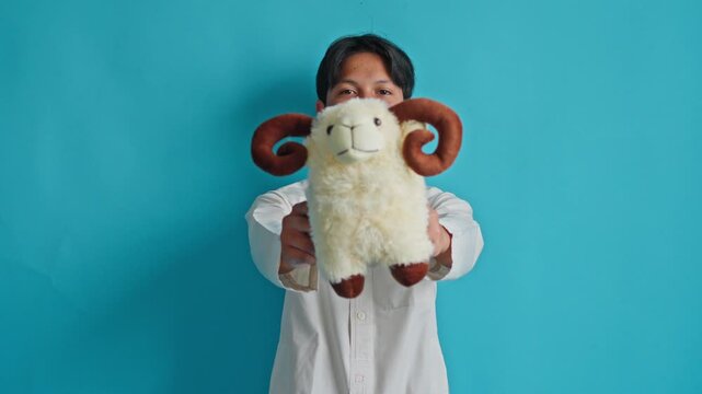 Smiling Asian teenager in robe shows plush sheep toy to camera for celebrating Eid al-Adha on blue background