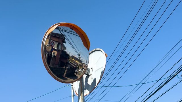 A convex traffic mirror reflecting a japanese town with telephone poles and wires under a clear blue sky on a sunny day in Japan.