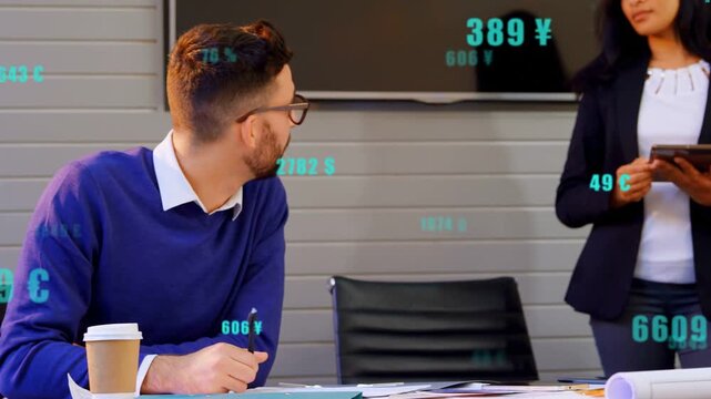 Seated man writing turning, standing woman tapping tablet showing currency data for finance review
