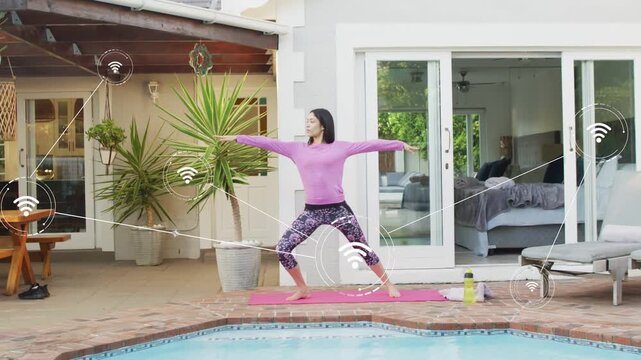 Woman stepping onto pink mat and holding Warrior II beside pool, wifi icons showing fitness data