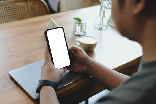 An over the shoulder view of a person holding a smartphone with a blank white mockup screen on a wooden cafe table next to a cup of coffee.