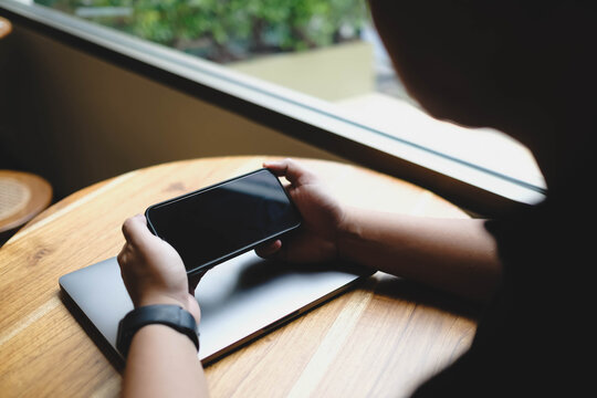 An over the shoulder view of a person holding a smartphone horizontally with a blank black mockup screen above a laptop on a wooden table.