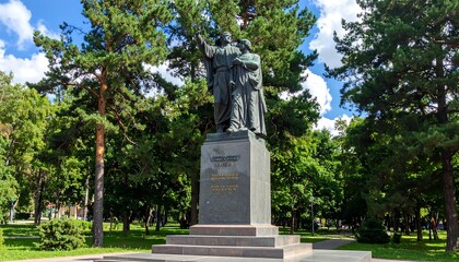 Fototapeta premium A large bronze sculpture of two figures stands atop a pedestal in a park, surrounded by lush green trees and a blue sky