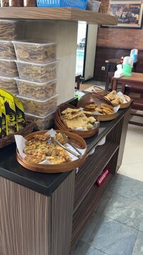 a self-serve Indonesian snack counter featuring fried favorites like bakwan, tempe, and tahu. traditional rattan trays with tongs sit beside stacked containers of crispy rempeyek crackers