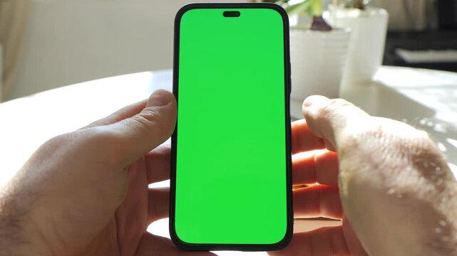 Hands hold smartphone with bright green screen. Phone centered in frame. Background shows table and white pots with plants. Sunlight creates soft shadows. Hands steady the phone without moving