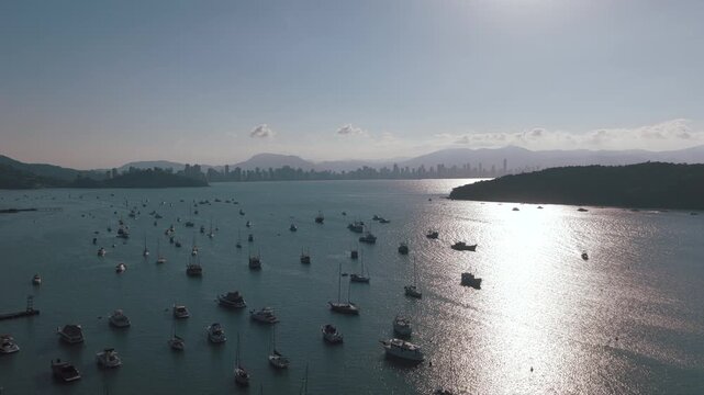 Anchored boats in Porto Belo bay Santa Catarina Brazil with Itapema and Meia Praia skyline backed by Serra do Mar coastal mountains under bright reflection on water, drone forward glide shot