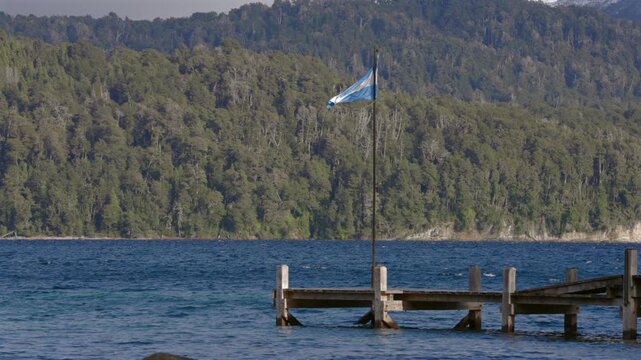 Argentine flag waving on a wooden pier over a deep blue lake surrounded by forest in Patagonia Argentina.