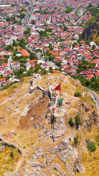 Vertical video. Fethiye, Turkey. Large Turkish flag flying on flagpole atop the historic Fethiye Castle hill with ancient Lycian rock cut tombs in the surrounding cliffs, drone footage.. Aerial View,