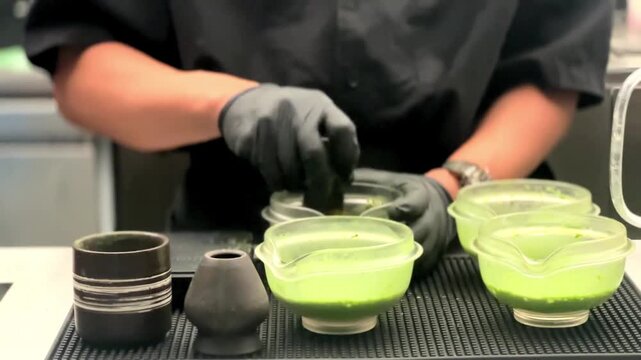 Professional hands prepare matcha with care and precision. Green tea is stirred in clear bowls with a traditional brush. Black gloves and dark apron suggest a focused, clean environment.