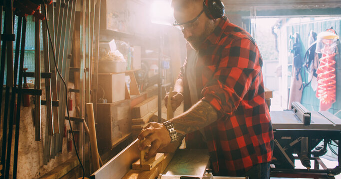 Joiner process. Furniture making. Man with ear protectors smoothing wood on thickness planer. Saw dust blur light in industry interior.