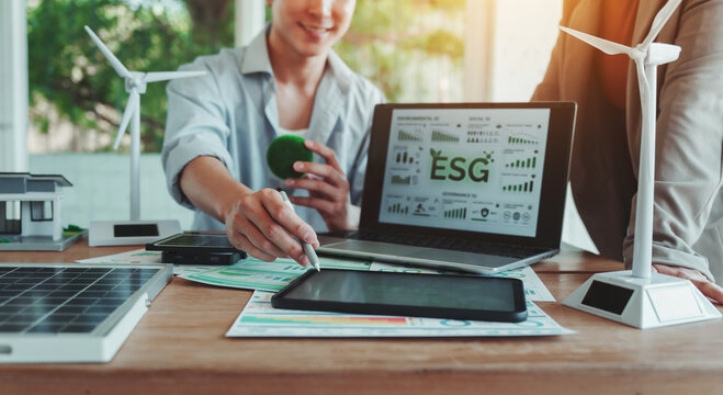Business team reviewing ESG sustainability report on laptop with solar panels, wind turbines and tablet on desk, renewable energy planning in modern office.