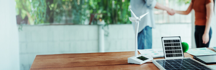 Renewable energy business meeting: tabletop model wind turbine and solar panels with charts, while two people shake hands in a bright office, suggesting clean power partnership. © Phushutter