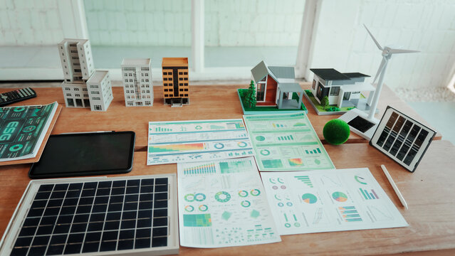 Desk workspace with solar panel, wind turbine model, smart home miniatures and printed energy analytics charts, illustrating renewable energy planning and sustainable city development.