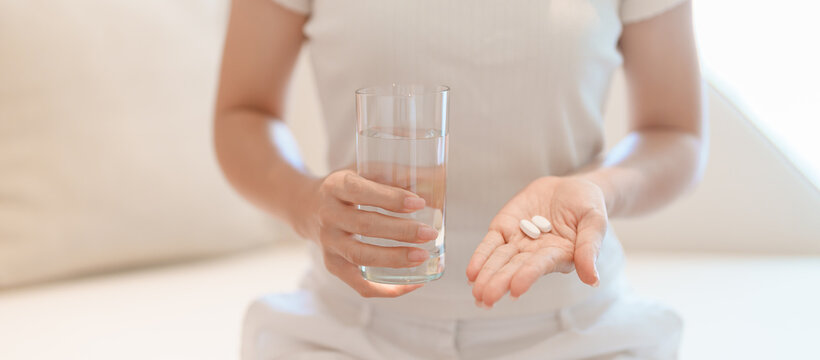 woman hand holding medicine painkiller pill and water glass on the sofa at home, taking for headaches, stomach ache, Diarrhea Pain from food poisoning, Endometriosis, Hysterectomy and Menstrual