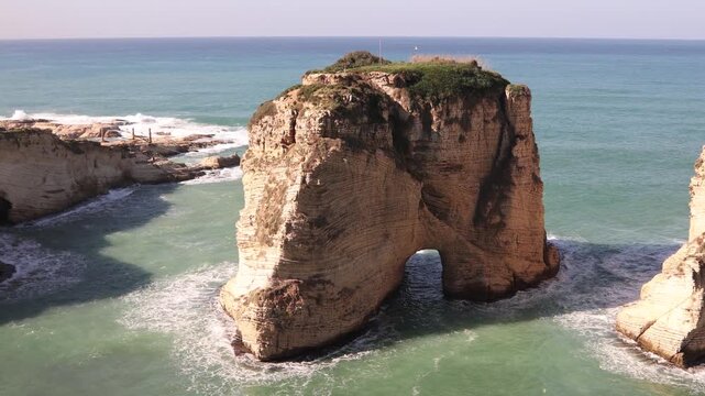 Panoramic view of the Raouche Pigeons Rock in Beirut. The shot captures the massive limestone arch and cliffs being hit by the turquoise Mediterranean waves, showcasing Lebanon