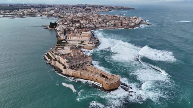 Aerial drone orbital shot of Maniace Castle at the tip of Ortigia Island, Syracuse. Medieval fortress architecture surrounded by the blue Mediterranean Sea and crashing waves in Sicily