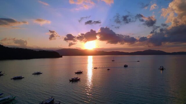 Traditional philipines outrigger bancas resting in a tranquil bay during an impressive colorful sunset, creating a peaceful tropical scene. Smooth aerial view descending panorama drone