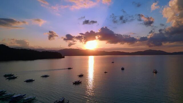 Tropical sun setting behind islands, reflecting golden light on peaceful water with outrigger bangka boats anchored. Magic aerial view drone shot panorama from above