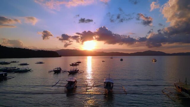 Traditional philipines outrigger bancas resting in a tranquil bay during an impressive colorful sunset, creating a peaceful tropical scene. Best aerial view ascending raise up panorama drone