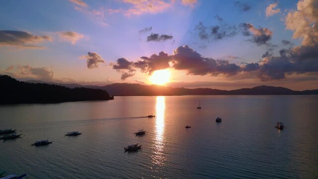 Tropical sun setting behind islands, reflecting golden light on peaceful water with outrigger bangka boats anchored. Nice aerial view overflight flyover panorama drone