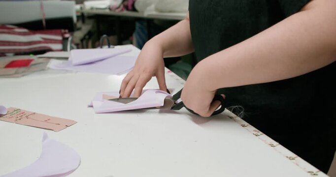 Sewing workshop. Clothing production. Female seamstress cutting white fabric with scissors figure paper pattern draft on studio desk.