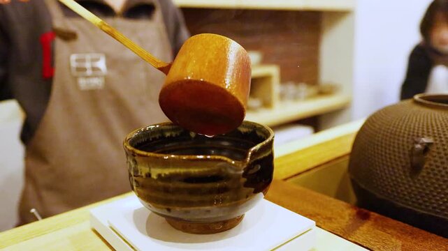 Close-up cinematic motion of hot water being poured from wooden ladle into traditional japanese tea bowl, highlighting steam and careful pouring technique in food and drink preparation.