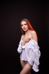 A young woman poses in a white off-the-shoulder shirt on a black background. The soft lighting highlights her relaxed and confident expression. She is wearing minimal underwear, which adds to her sens © SergeiPh