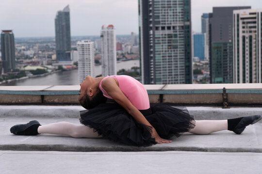 Flexible young Asian girl ballerina in pink leotard and black tutu practicing a split and backbend on rooftop with city skyline background. Urban gymnastics and dancing lifestyle concept.