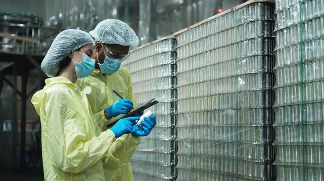 Two industrial engineer supervisors inspect and examine quality of stainless steel containers, production process in canned food manufacturing factory, and protective uniforms for hygiene operations.