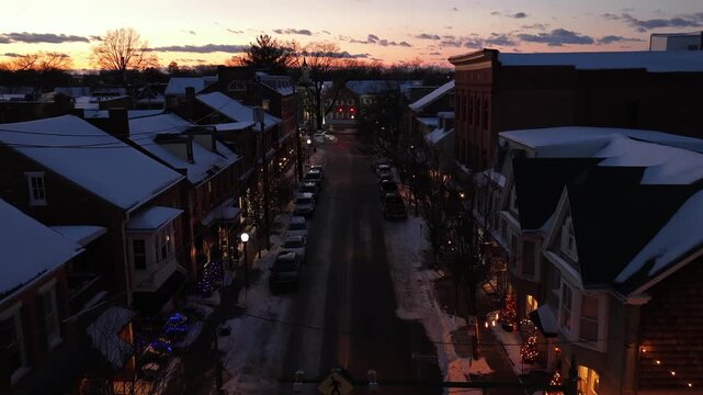 Historic small-town street at winter twilight with snow-covered rooftops, warm lantern lights and cozy two-story buildings. Charming and peaceful evening atmosphere. Aerial flyover shot.