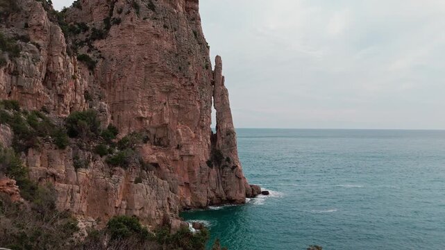 A stunning cinematic aerial shot of the iconic Pedra Longa limestone needle rising from the turquoise Mediterranean sea in Baunei, Sardinia. Great for travel and nature documentaries.