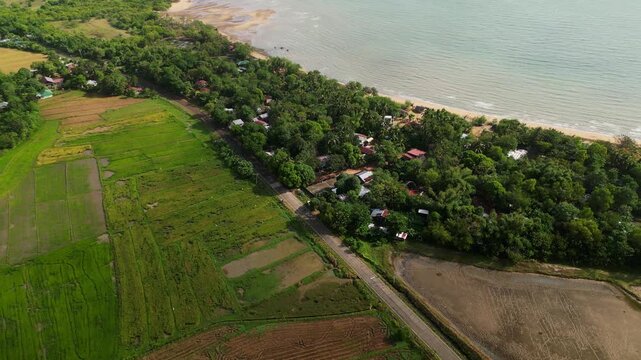 Aerial flyover of a coastal road passing through lush tropical greenery, farmland, and village homes along the shoreline in Guimaras Island, Philippines