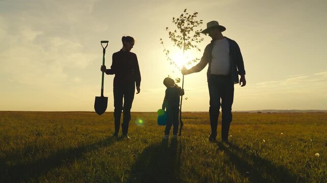 A family of three is walking through a field, with a man holding a shovel and a child holding a tree sapling. The scene is peaceful and serene, with the sun setting in the background
