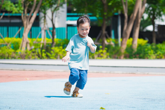 A happy young Asian boy in a light blue t-shirt runs across a blue playground surface in a sunny public park. Concept of childhood energy, active lifestyle, and physical development for kids.