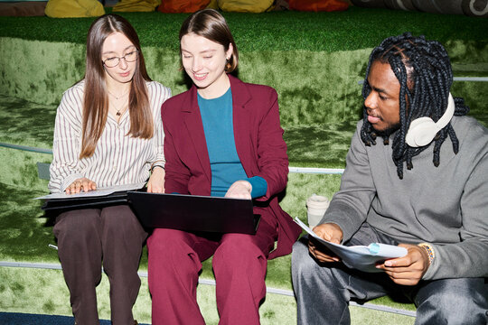 Multiethnic young coworkers reviewing laptop and documents while collaborating on steps, representing teamwork and planning. Useful for business communication, startup marketing, recruitment
