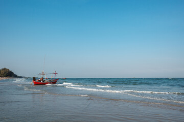 Obraz premium A serene, wide-angle shot of a traditional, brightly colored red fishing boat anchored in the gentle surf of a calm, sandy beach. The clear blue sky meets the horizon over the tranquil sea.