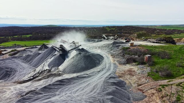 Machines are working at a granite quarry site. Stone is being crushed and stockpiled in large mounds. Dust is rising into the air as workers manage the process.