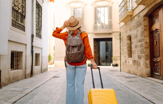 A woman walks along a narrow street, pulling a bright yellow suitcase. She wears a backpack and a wide-brimmed hat. Sunlight shines on the buildings around her.