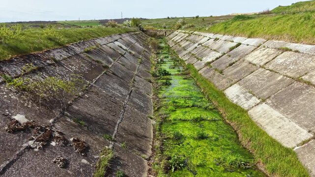 Water channels are dry, with patches of green plants growing in the center. Grass surrounds the edges of the concrete structure under a clear sky. The land shows signs of water absence.