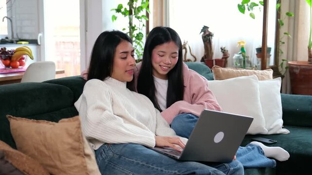 Two happy girlfriends using a laptop for online shopping while sitting on a couch at home