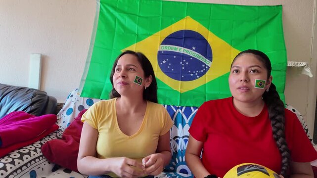 Two Latina women with Brazilian flags painted on their faces watching a soccer championship at home