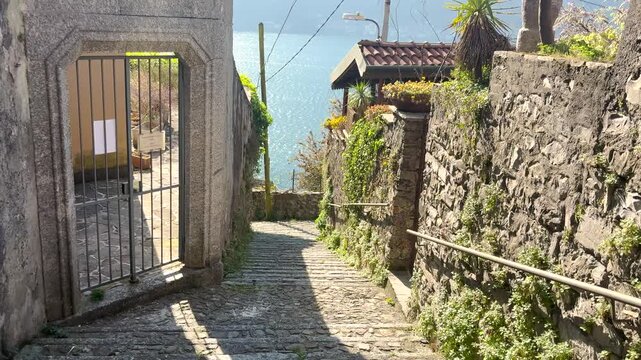 POV walking through narrow stone streets of Nesso with blue lake views, picturesque Italian village with rustic architecture and coastal atmosphere on Lake Como