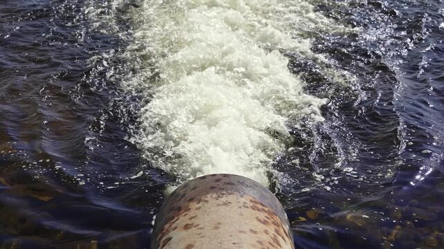 Bright daylight illuminates a relentless gush of foamy water from an old, mottled pipe. It violently enters the dark, deep river, generating captivating ripples and a strong current.