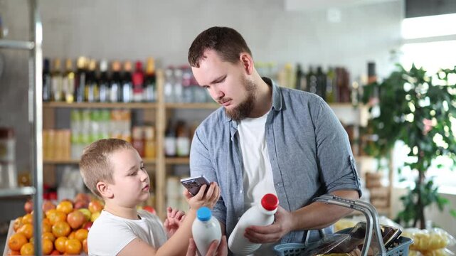 Focused tween boy with friendly young father scanning barcodes on bottles with milk with phone while shopping in supermarket, checking product information and price 